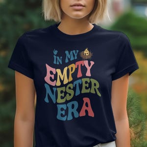 May include: A navy blue t-shirt with the text "In My Empty Nester Era" in colorful letters. The text is stylized with a retro font and includes a small bird and a birdhouse graphic.