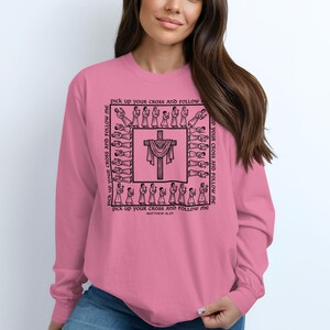 May include: A long-sleeved, pink sweatshirt featuring a black graphic of a cross with a draped cloth, surrounded by figures. The text "Pick up your cross and follow me" is printed around the cross and "Matthew 16:24" is printed at the bottom.