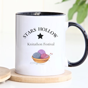 Könnte beinhalten: Weiße Keramiktasse mit schwarzem Rand und Henkel. Die Tasse zeigt einen schwarzen Stern und den Text "Stars Hollow Knitathon Festival". Unter dem Text ist ein Korb mit Garn und Stricknadeln abgebildet.