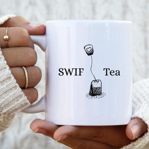 Könnte beinhalten: Ein weißer Keramikbecher mit einer schwarzen Grafik eines Teebeutels mit einem Herz auf dem Etikett und dem Text SWIF Tea in schwarzen Buchstaben. Das Design ist auf der Vorderseite des Bechers zentriert, der eine glatte Oberfläche hat.