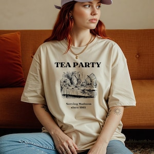 Könnte beinhalten: Beigefarbenes T-Shirt mit dem Schriftzug "TEA PARTY" über einer Schwarz-Weiß-Illustration einer Teeparty-Szene. Darunter steht der Text "Serving Madness since 1865". Das Shirt hat eine lockere, übergroße Passform.