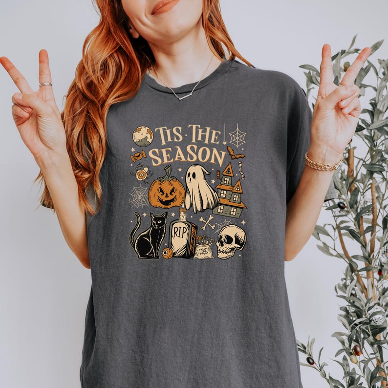 May include: Dark gray t-shirt with a Halloween-themed graphic. The design includes a pumpkin, ghost, haunted house, black cat, tombstone, and skull. The text "Tis The Season" is printed above the illustrations.
