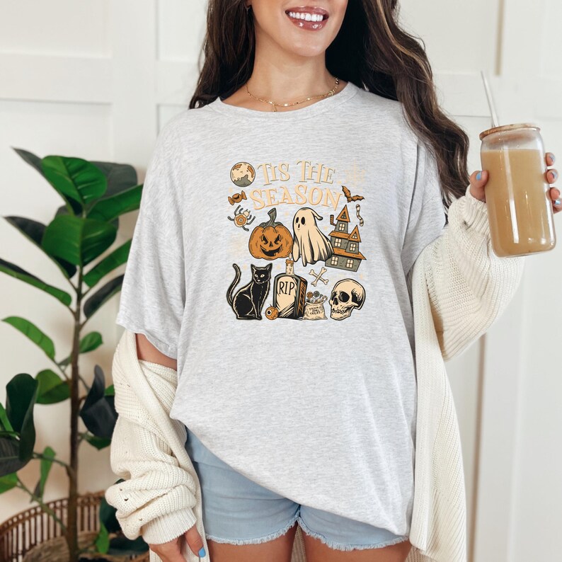 May include: A light gray t-shirt with a Halloween-themed graphic. The graphic includes a jack-o'-lantern, ghost, haunted house, black cat, skull, and the text "'Tis The Season". The shirt is paired with blue shorts and a cream-colored cardigan.