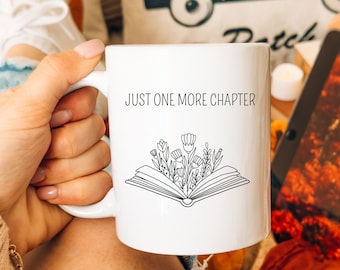 May include: A white ceramic mug featuring a black line art illustration of an open book with flowers growing out of the pages. The text "JUST ONE MORE CHAPTER" is printed in a simple black sans-serif font above the book graphic on the mug.
