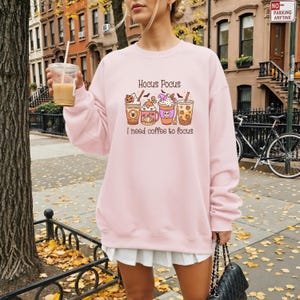 Może przedstawiać: Jasnoróżowa bluza z napisem "Hocus Pocus" nad czterema ilustrowanymi filiżankami kawy z motywami Halloween. Poniżej filiżanek znajduje się napis "I need coffee to focus". Bluza ma okrągły dekolt.
