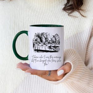 Könnte beinhalten: Weißer Keramikbecher mit grünem Henkel und Rand. Der Becher zeigt eine Schwarz-Weiß-Illustration einer Szene aus Alice im Wunderland mit dem Zitat "Ich wusste, wer ich heute Morgen war, aber ich habe mich seitdem ein paar Mal verändert."