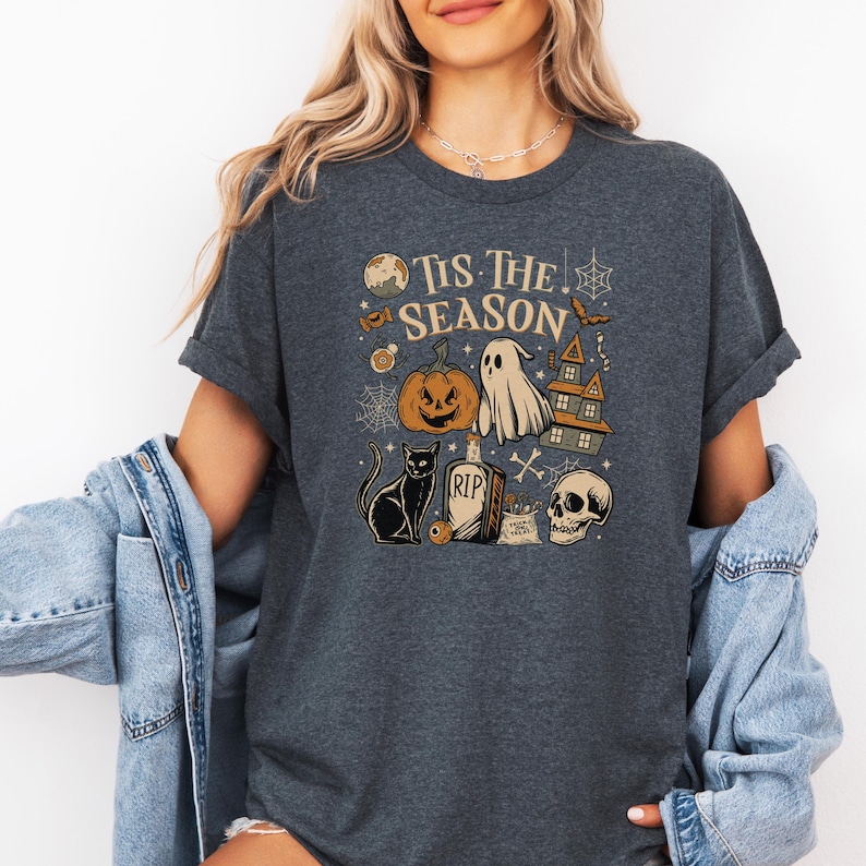 May include: Dark gray t-shirt with a Halloween-themed graphic. The design includes a jack-o'-lantern, ghost, haunted house, black cat, skull, and the text "Tis The Season". The shirt has short sleeves and a relaxed fit.