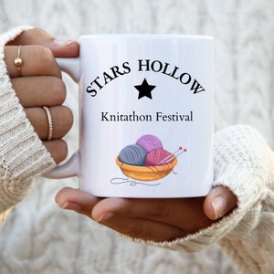Könnte beinhalten: Ein weißer Keramikbecher mit dem schwarzen Schriftzug Stars Hollow, einem schwarzen Sternsymbol und dem Text Knitathon Festival. Das Design zeigt eine Grafik einer Holzschale, die mit drei Wollknäueln in Grau, Lila und Pink gefüllt ist.