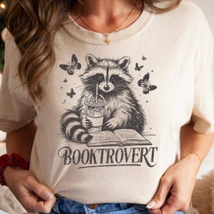 Könnte beinhalten: Cremefarbenes T-Shirt mit einer detaillierten Schwarz-Weiß-Illustration eines Waschbären, der aus einem Getränk trinkt, mit Schmetterlingen und einem offenen Buch. Der Text "BOOKTROVERT" ist unter dem Waschbären aufgedruckt.