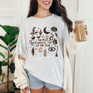 Könnte beinhalten: Ein graues T-Shirt mit einem schwarzen und orangefarbenen Druck verschiedener Halloween-Symbole, darunter ein Hexenhut, ein Halbmond, ein Kürbis, ein Schädel, eine Zaubertrankflasche, ein Auge, eine Feder und ein Besen. Der Text "It's The Most Wonderful Time Of The Year" ist auf das T-Shirt gedruckt.