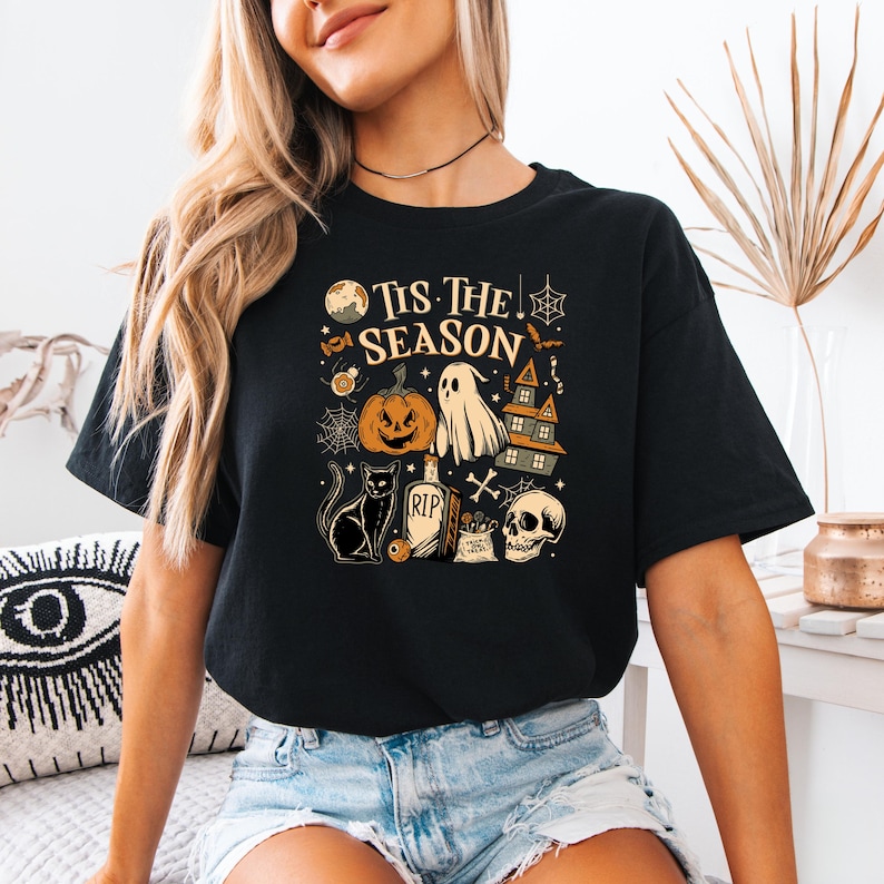 May include: Black t-shirt with a Halloween-themed graphic. The design includes a jack-o'-lantern, ghost, haunted house, black cat, skull, and the text "Tis The Season". The shirt is made of a soft material.