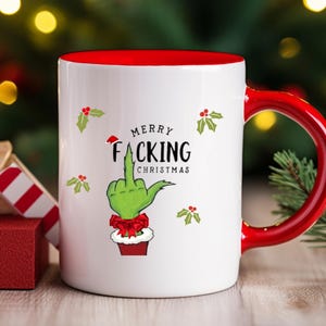 Könnte beinhalten: Ein weißer Keramikbecher mit leuchtend rotem Innenraum und Henkel. Auf der Vorderseite ist eine grüne Hand zu sehen, die den Mittelfinger zeigt, mit einer roten Schleife und dem Text Merry Facking Christmas sowie kleinen grünen Stechpalmenblättern.