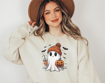 May include: A cream-colored crewneck sweatshirt featuring a graphic of a white ghost wearing an orange knit beanie. The ghost holds an iced coffee and a jack-o'-lantern pumpkin, surrounded by small black bats and stars against a plain background.
