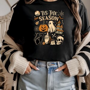 May include: Black t-shirt with a Halloween-themed graphic. The design includes a jack-o'-lantern, ghost, haunted house, black cat, tombstone, skull, and the text "Tis The Season". The graphic is in shades of orange, white, and black.