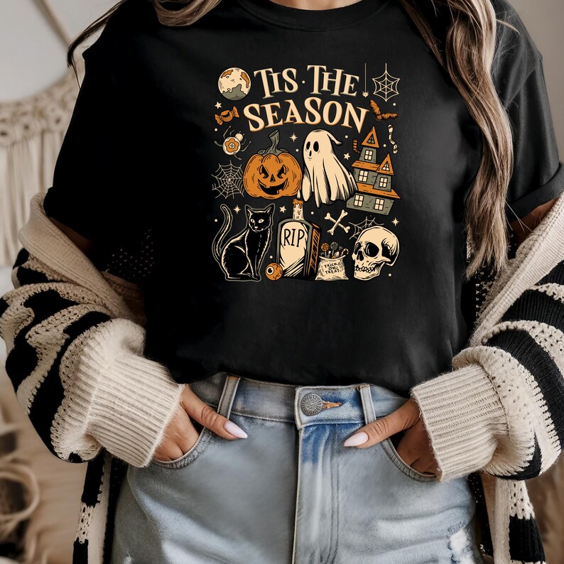 May include: Black t-shirt with a Halloween-themed graphic. The design includes a jack-o'-lantern, ghost, haunted house, black cat, tombstone, skull, and the text "Tis The Season". The graphic is in shades of orange, white, and black.