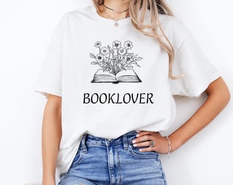 Peut inclure: T-shirt blanc avec une illustration en noir et blanc d'un livre avec des fleurs qui en sortent. Le texte "BOOKLOVER" est imprimé sous l'illustration.