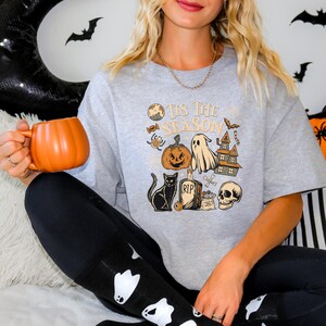 May include: A light gray sweatshirt with a Halloween-themed graphic that includes a pumpkin, ghost, black cat, skull, and the text "It's The Season." The person is holding an orange pumpkin-shaped mug.