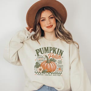 Könnte beinhalten: Ein cremefarbenes Sweatshirt mit Rundhalsausschnitt und einem Grafikdruck eines orangefarbenen Kürbisses mit grünen Blättern und Blüten. Der Text lautet PUMPKIN PATCH, FRESH FROM THE PATCH, FARM FRESH, LOCALLY GROWN, AUTUMN PICKED, PUMPKINS, CORN MAZE, APPLE CIDER.