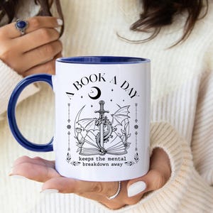 Pode incluir: Uma caneca de cerâmica branca com interior e alça azul-escura. A caneca apresenta um gráfico de linhas pretas de um dragão enrolado em uma espada sobre um livro aberto, com o texto A BOOK A DAY keeps the mental breakdown away.