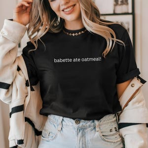 Puede incluir: Una camiseta negra de manga corta con el texto blanco "babette ate oatmeal!" impreso en el pecho. La camiseta tiene cuello redondo y se lleva con una chaqueta blanca con rayas negras y unos vaqueros de color azul claro.