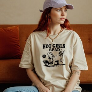 Könnte beinhalten: Beiges T-Shirt mit dem Text "HOT GIRLS READ" und einer Schwarz-Weiß-Illustration einer lesenden Frau im Vintage-Stil. Das T-Shirt hat eine lockere, übergroße Passform.