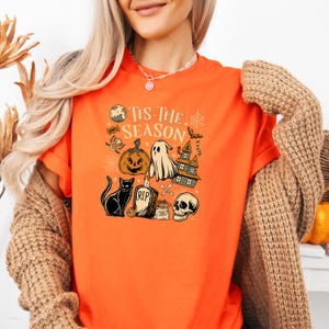 May include: Orange t-shirt with a Halloween-themed graphic. The design includes a jack-o'-lantern, ghost, haunted house, black cat, tombstone, skull, and the text "Tis The Season". The shirt is a short-sleeved crew neck.