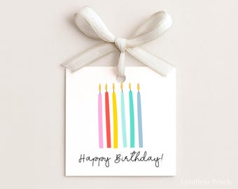May include: A white gift tag with a cream-colored ribbon bow. The tag features six colorful candles with yellow flames and the words "Happy Birthday!" in black script. The candles are pink, red, yellow, teal, turquoise, and blue.