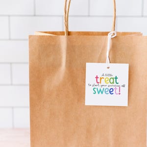 May include: A brown paper gift bag with twisted paper handles. A white tag is attached with the text "A little treat to start your summer off sweet!" in colorful lettering. The background is a white tile wall.