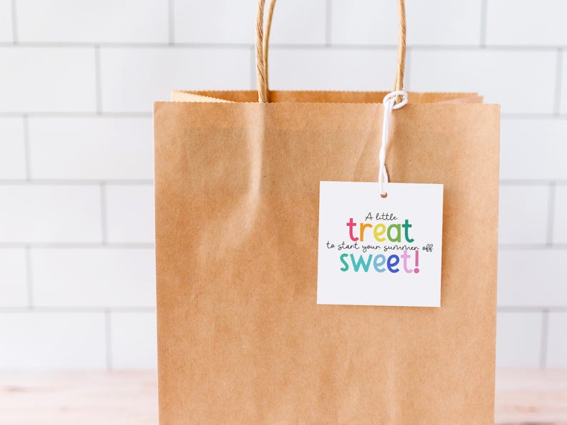 May include: A brown paper gift bag with twisted paper handles. A white tag is attached with the text "A little treat to start your summer off sweet!" in colorful lettering. The background is a white tile wall.