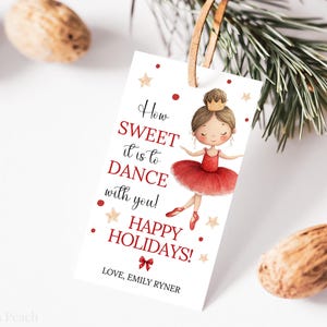 May include: A white gift tag with a watercolor illustration of a ballerina in a red tutu. The tag includes the text "How SWEET it is to DANCE with you! HAPPY HOLIDAYS!" and is tied with a brown leather cord.