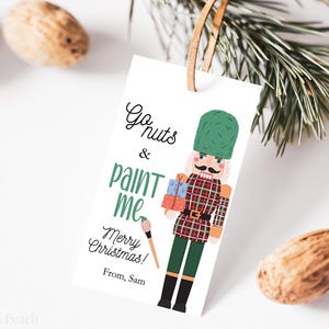 May include: A white gift tag with a cartoon nutcracker illustration. The tag reads "Go nuts & paint me Merry Christmas! From, Sam." The nutcracker wears a green hat and a plaid jacket. Brown nuts and pine branches are in the background.