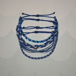 May include: A set of blue and white friendship bracelets with a silver turtle charm. The bracelets are made of braided string and beads.