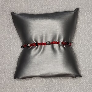May include: A red, white, and black beaded bracelet with a silver charm. The bracelet is lying on a gray pillow.