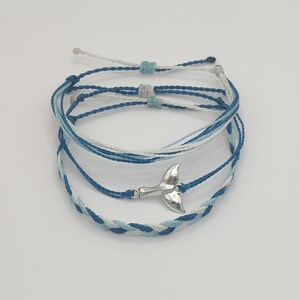 May include: Set of four blue and white braided friendship bracelets with a silver whale tail charm.
