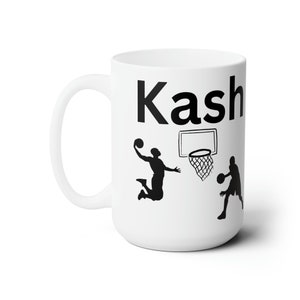 Könnte beinhalten: Weiße Keramiktasse mit einer schwarzen Silhouette von zwei Basketballspielern und einem Basketballkorb. Die Tasse trägt den Namen "Kasha" aufgedruckt.