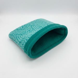 May include: A small, rectangular pouch featuring a soft, teal-colored fleece interior and a textured exterior with a light teal and white herringbone-style knit pattern. The pouch has a wide, open top with a thick, folded-over teal fleece edge.