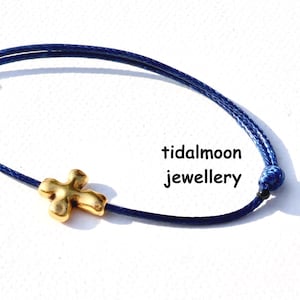 May include: A navy blue cord bracelet with a gold-coloured cross charm. The bracelet has a sliding knot closure and the text "tidalmoon jewellery". The jewellery is set against a white background.