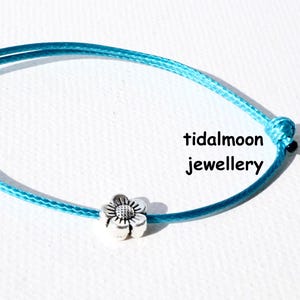 May include: A turquoise adjustable bracelet with a silver flower charm. The bracelet is made of a thin, woven cord and features a sliding knot closure. The text "tidalmoon jewellery" is visible.