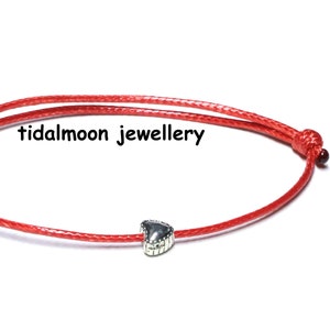 May include: A red string bracelet with a silver heart charm. The bracelet is tied with a knot at each end. "tidalmoon jewellery" is printed on the white background.