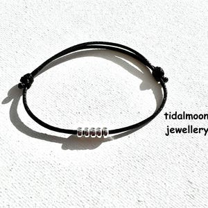 May include: Black string bracelet featuring five silver beads. The bracelet is knotted at each end for adjustment. The text "tidalmoon jewellery" is visible. The bracelet is on a white textured surface.