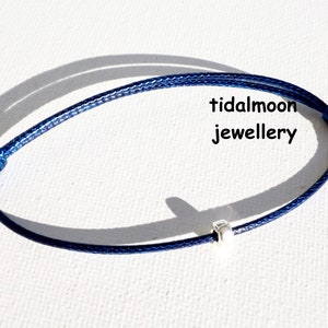 May include: A navy blue cord bracelet with a silver bead charm. The bracelet is tied with a knot at each end. The text "tidalmoon jewellery" is visible in the image.