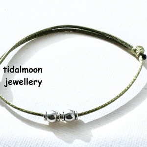 May include: A green string bracelet with two silver beads. The bracelet is tied with a knot and has the text "tidalmoon jewellery" on the white background.