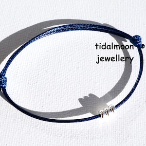May include: A navy blue adjustable bracelet with three small silver beads. The bracelet is made of a woven cord and features the text "tidalmoon jewellery". The bracelet is displayed on a white surface.