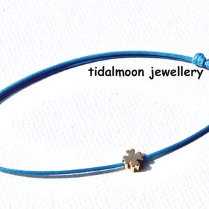 May include: A blue cord bracelet featuring a gold-coloured four-leaf clover charm. The image includes the text "tidalmoon jewellery". The bracelet is knotted at both ends.
