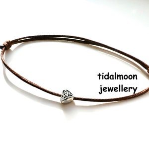 May include: A brown cord bracelet with a silver Celtic knot heart charm. The bracelet is tied with a simple knot and the charm is centred on the cord. The text "tidalmoon jewellery" is visible in the image.
