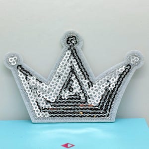 May include: A silver sequined crown patch with a grey felt backing. The crown features a classic five-point design with a silver sequin pattern and black accents. The patch is suitable for embellishing clothing or accessories.