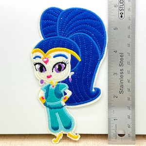 May include: An embroidered patch depicting a cartoon genie with blue hair, purple eyes, and a teal outfit. The patch is about 7.6 cm tall and is made of fabric with a stitched design. The patch is on a white background.