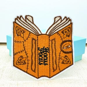 Könnte beinhalten: Gestickter Aufnäher mit einem offenen Buch mit dem Text "Hocus Pocus" in Schwarz. Das Buch hat einen orangefarbenen Einband mit braunen Akzenten und dekorativen Nähten. Der Aufnäher ist weiß umrandet.