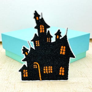 May include: Black embroidered patch of a haunted house with orange lit windows and a street lamp. The patch has a white border and is set against a light blue box, perfect for Halloween-themed crafts and apparel.