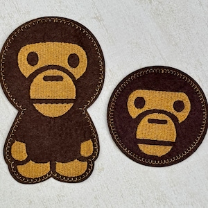 Peut inclure: Deux patchs en feutre marron avec des images brodées d'un singe. Le plus grand patch a un singe avec des bras et des jambes, tandis que le plus petit patch a seulement une tête de singe.